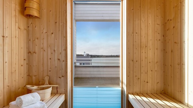 A sauna room with wooden walls, a bench, a towel stack, and a window with a view of buildings and a body of water. - Cozy home in Olpenitz with sauna (Olpenitz)