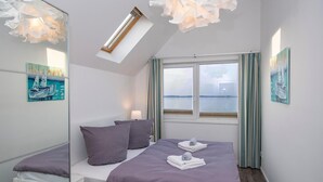 Ein Schlafzimmer mit Bett, Kissen, Nachttisch, Lampe, einem Fenster mit Vorhängen und Bildern an den Wänden.