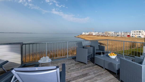 Ein Balkon mit Korbmöbeln, einem kleinen Tisch mit Obstschale und Blick auf das Meer und Gebäude in der Ferne.