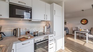 Moderni keittiö, jossa on valkoiset kaapit, Bosch-merkkinen mikroaaltouuni ja uuni. Keittiössä on myös ruokailutila, jossa on pöytä ja tuolit, ja ikkunasta avautuu näkymä auringonlaskuun.