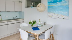 A modern kitchen with white cabinets, a dining area with a white table and chairs, a decorative light, and a beach-themed painting on the wall. - Lovely apartment in Olpenitz with sauna (Olpenitz)