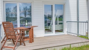 A wooden deck with a table and chairs, a glass door leading to a parking area, and a metal railing. - Lovely apartment in Olpenitz with sauna (Olpenitz)