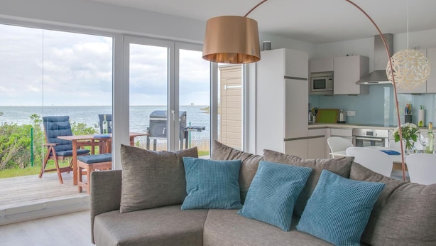 A modern living room with a grey sofa, blue cushions, and a view of the sea through large windows. - Lovely apartment in Olpenitz with sauna (Olpenitz)