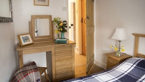 A bedroom with a wooden desk, a bed with a plaid blanket, a nightstand with a lamp, and a door leading to another room.