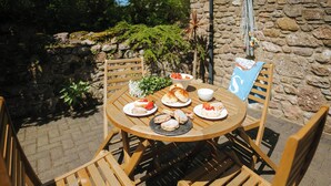 A round wooden table with plates of food, surrounded by wooden chairs, set outdoors with a stone wall and greenery in the background. - 2 bedroom accommodation in Gower (Gower)