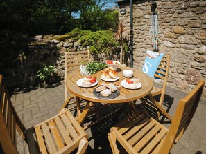 A round wooden table with plates of food, surrounded by wooden chairs, set outdoors with a stone wall and greenery in the background.