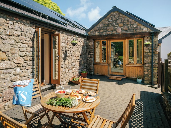A stone building with wooden doors and windows, a round wooden table with plates of food, and a bench.