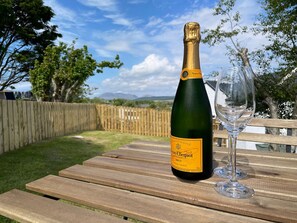 Een fles champagne en een glas op een houten tafel, buiten met een houten hek en bomen op de achtergrond.