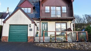 A two-story house with a green garage door, a balcony with a railing, and a stone wall in front. - 3 bedroom accommodation in Lamlash (Lamlash)