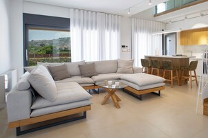 Una sala moderna con un sofá seccional, una mesa de centro de cristal y un comedor con sillas de madera, con una cocina al fondo.