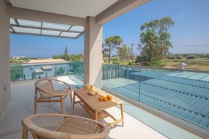 Un balcón con barandilla de vidrio, dos sillas de mimbre, una mesa de madera con naranjas y vasos, y vistas a una piscina y zonas verdes.