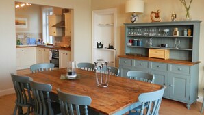 Zona pranzo con tavolo di legno e sedie blu, una cucina sullo sfondo e un armadietto blu con bicchieri.