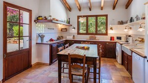 A kitchen with wooden beams, a dining table, chairs, a washing machine, and a window with a view of greenery. - VILLA CAN TEULADA (Puerto Pollensa)