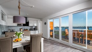 A modern kitchen with white cabinets, a dining area with a table set for two, and a large sliding glass door leading to an outdoor deck with a view of the water. - Beautiful apartment in Olpenitz (Olpenitz)