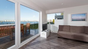 A modern living room with a large sliding glass door leading to a balcony with a view of the sea and beach houses. - Beautiful apartment in Olpenitz (Olpenitz)