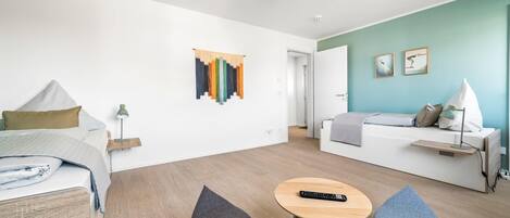 Een moderne hotelkamer met een bed, een slaapbank, een kleine tafel en een stoel. Er hangt een kunstwerk aan de muur en een deur naar een andere kamer.