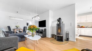 A modern living room with a sofa, coffee table, and yellow cushions. A fireplace is visible in the background, and a kitchen area is partially seen. - Lovely apartment in Olpenitz with sauna (Olpenitz)