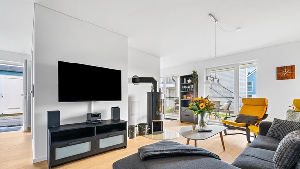 A modern living room with a flat-screen TV, a black wood-burning stove, a sofa, a yellow armchair, and a dining area with a vase of flowers. - Lovely apartment in Olpenitz with sauna (Olpenitz)
