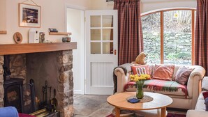 Una sala de estar acogedora con chimenea de piedra, un sofá cómodo, una mesa redonda de madera, y vistas a un muro de piedra y vegetación.