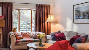 Una sala de estar acogedora con un sofá, una mesita de centro y una lámpara. Se ve una ventana con cortinas, un cuadro en la pared y vistas al exterior.