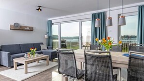 Un salón moderno con sofá, mesa de comedor, sillas y vista a una masa de agua a través de grandes ventanales.