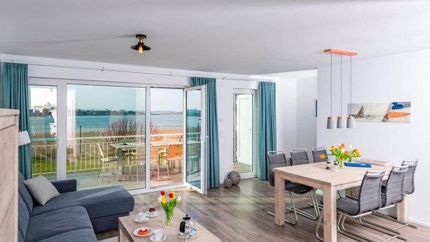 Una sala de estar moderna con un sofá, una mesa de comedor y sillas. Se ve el mar a través de grandes puertas de vidrio.