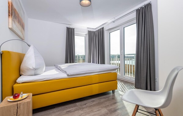 A modern hotel room with a yellow bed, a white chair, and large windows with grey curtains. - Nice home in Olpenitz with sauna (Olpenitz)