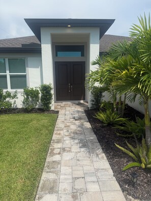 A modern house with a stone pathway leading to a double door, surrounded by greenery and a well-maintained lawn. - NEW Coastal Paradise GEM!! Spacious 4BR, 3 Full Baths w/Pool!! PET FRIENDLY!!! (Bonita Springs)