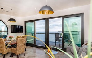 Quarto moderno com área de jantar, televisão e amplas janelas com vista para o mar.