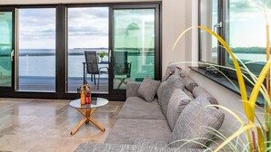 Un salon moderne avec un canapé gris, une table basse en verre et une vue sur la mer grâce à de grandes portes coulissantes.