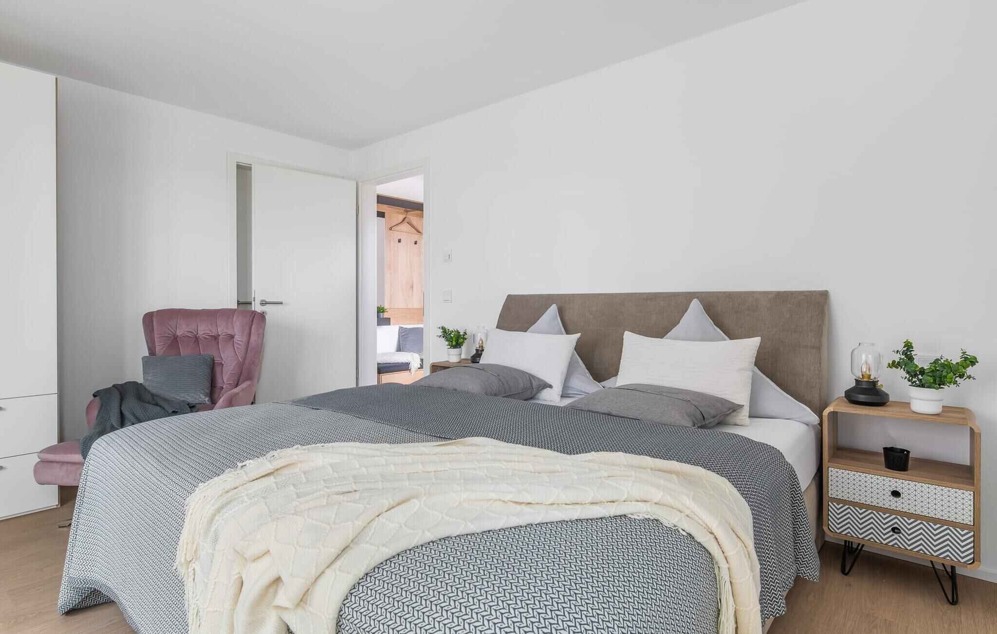 A modern bedroom with a large bed, a chair, a bedside table with a lamp, and a small plant.