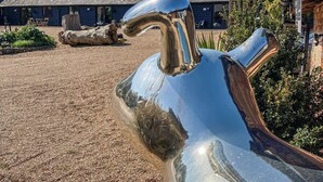 A large metallic sculpture in an outdoor setting with a gravel ground, a wooden structure in the background, and some greenery. - An amazing and extraordinary property. (Hastingleigh, near Ashford)