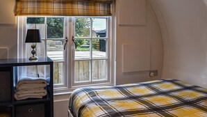 A bedroom with a plaid bedspread, a bedside lamp, a window with a plaid valance, and a view of a wooden deck and greenery outside. - An amazing and extraordinary property. (Hastingleigh, near Ashford)