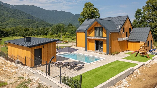 Una moderna cabina di legno con piscina, immersa in un prato curato e con montagne sullo sfondo.