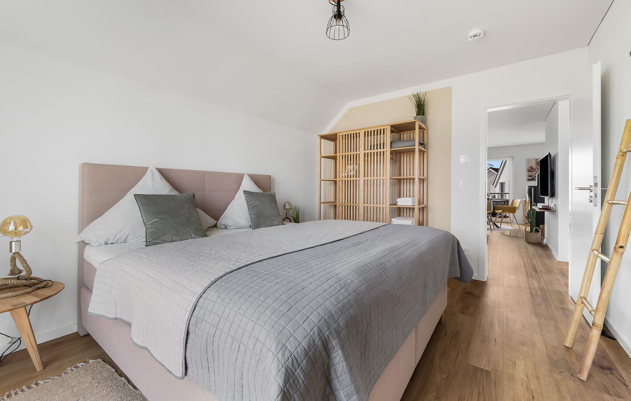 A bedroom with a large bed, bedside tables, a wooden wardrobe, and a view into a living area.