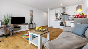 A modern living room with a flat-screen TV, a sofa, a coffee table with fruits, and a kitchen in the background. - Beautiful apartment in Olpenitz (Olpenitz)