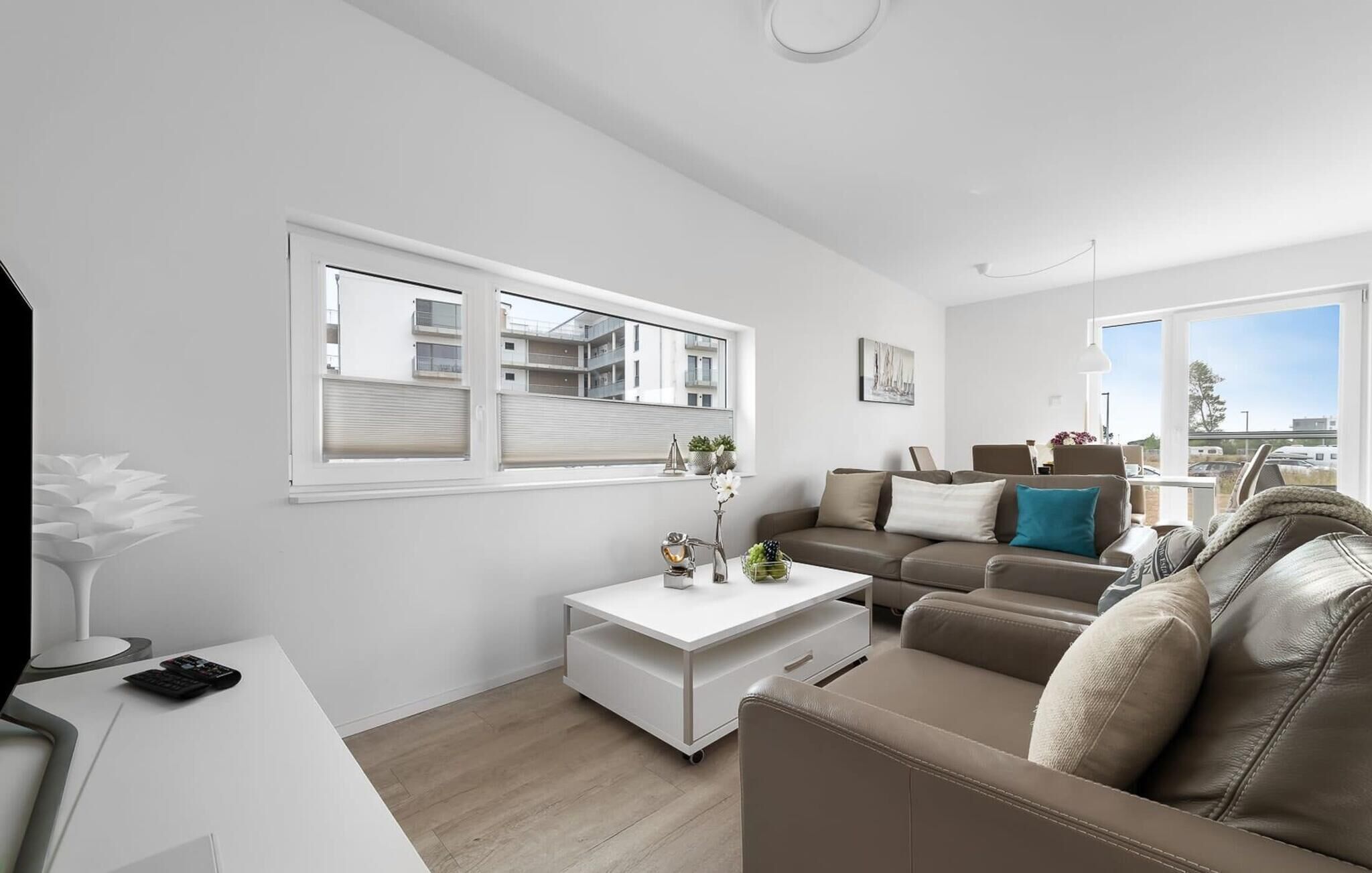 A modern living room with a white sofa, a coffee table, and a TV on a white stand. There are windows with blinds, a view of buildings outside, and a ceiling light.