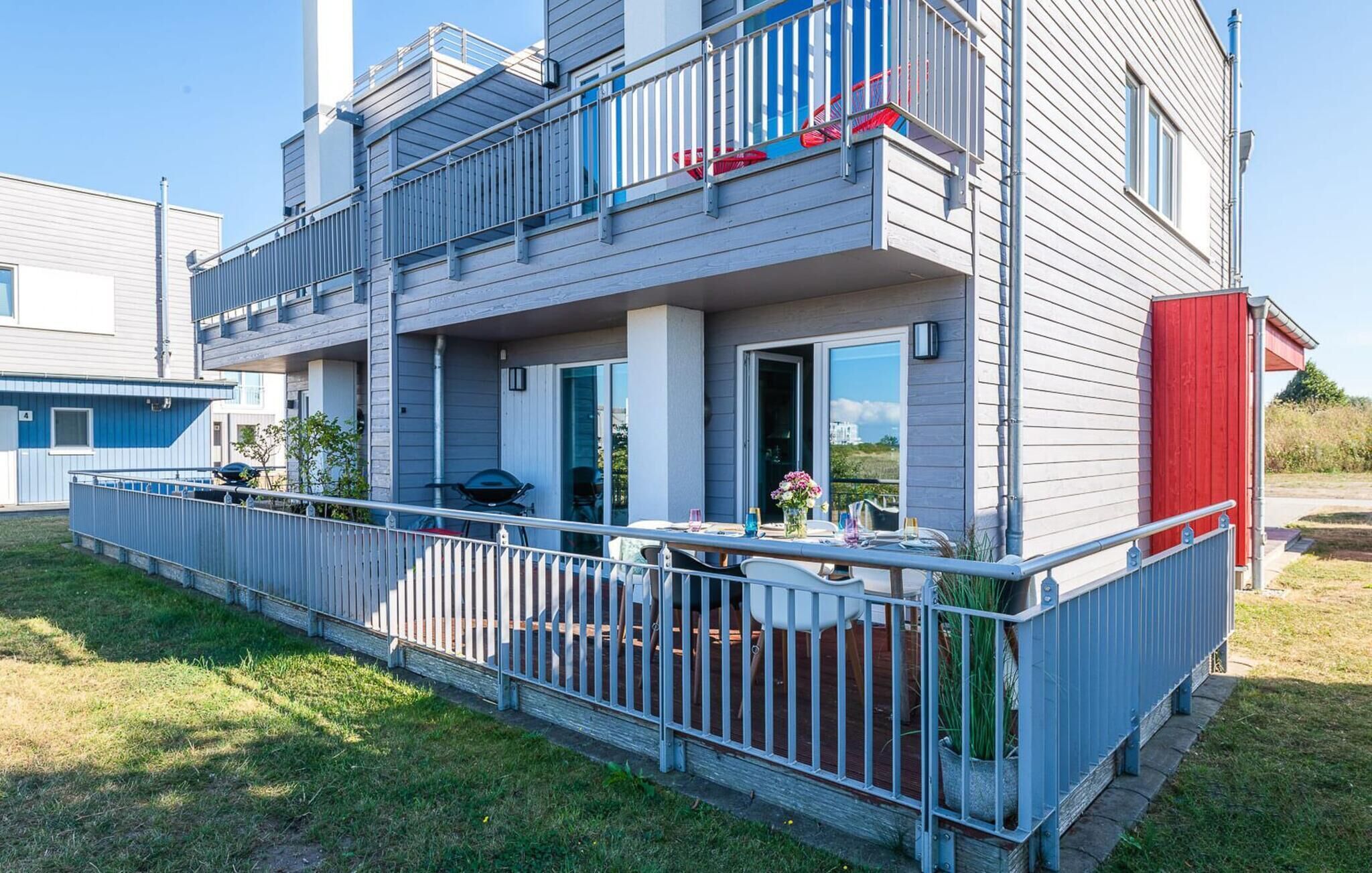 A modern two-story building with a balcony, a barbecue grill, and a red door.