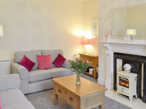 A living room with a grey sofa, red cushions, a wooden coffee table, a vase with flowers, a lamp, and a white fireplace with a clock above it. - 2 bedroom accommodation in Embleton, near Alnwick (Embleton, near Alnwick)