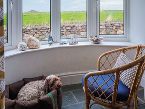 A room with a bay window overlooking a stone wall and green field. A wicker chair with a blue cushion and a dog bed with a dog are visible. - Chapel Cottage - UKC4683 (Clynnog Fawr)