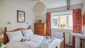 A bedroom with a wooden bed, a dresser, a window with curtains, a radiator, and a bedside table with a lamp. - Chapel Cottage - UKC4683 (Clynnog Fawr)