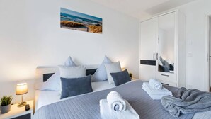 Ein modernes Hotelzimmer mit einem Bett, einem Nachttisch mit Lampe und Fernbedienung, einem großen Kleiderschrank und einem Poster mit Strandblick.