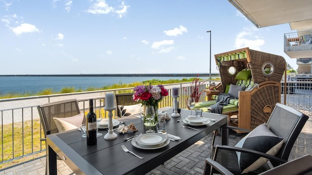 A dining table set with plates, cutlery, and a bottle of wine, overlooking a body of water with a bridge in the distance.
