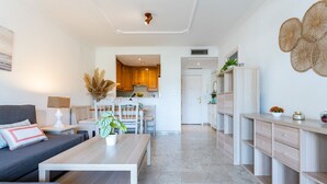 Un salon moderne avec un canapé, une table basse et des plantes décoratives. On aperçoit une cuisine à l’arrière-plan, avec des meubles en bois et un coin repas.