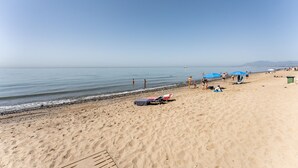 Une vaste plage de sable fin, un ciel dégagé, une mer calme et des montagnes à l’horizon.