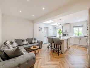 Uma sala de estar moderna com um sofá cinzento, uma mesa redonda pequena e uma área de cozinha com armários brancos e um padrão de piso em herringbone.