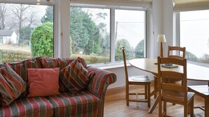 A cozy room with a striped sofa, a wooden dining table, and chairs. Large windows offer a view of greenery outside.