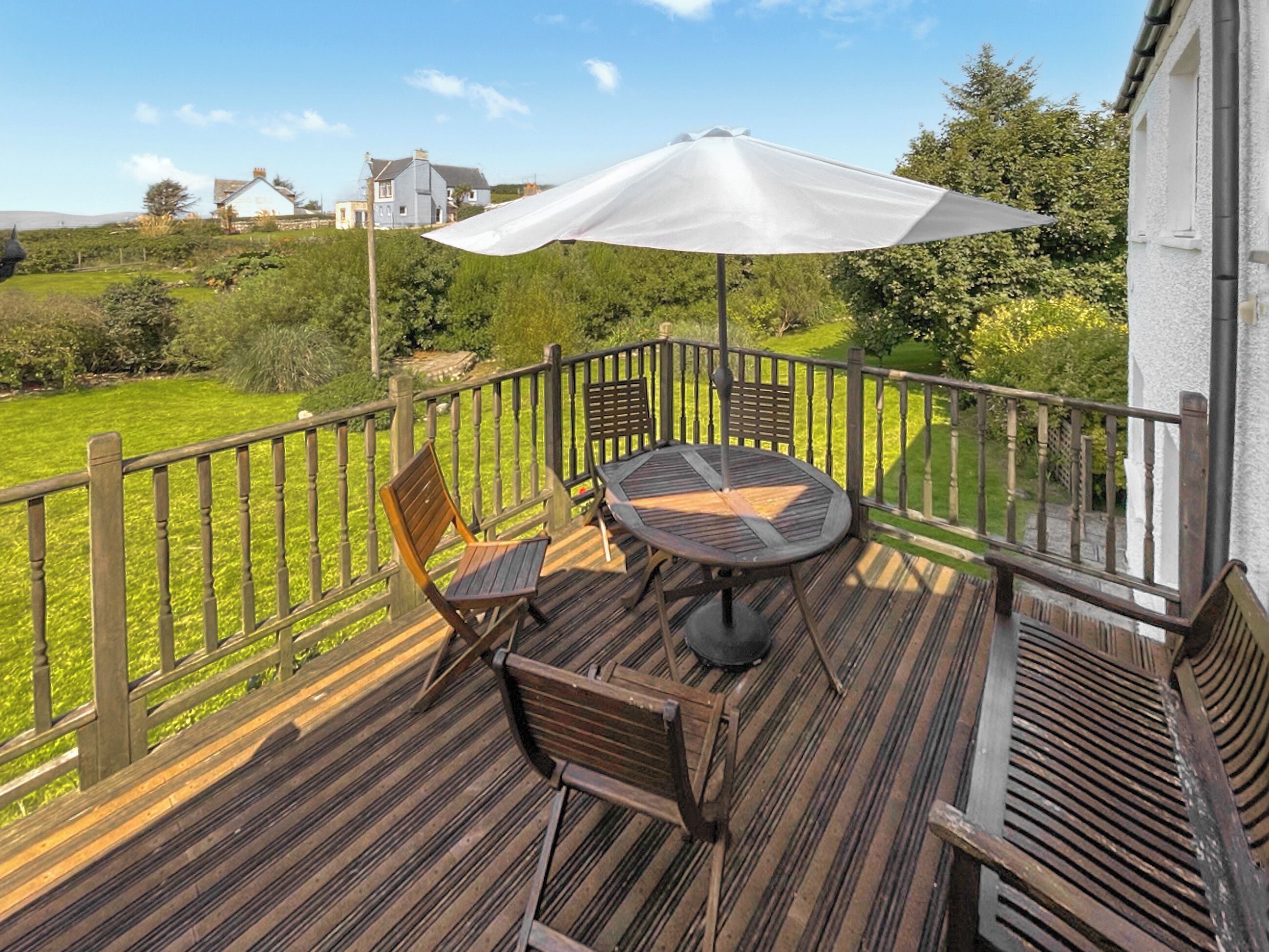 Eine Holzterrasse mit Tisch und Stühlen, einem weißen Sonnenschirm und Blick auf eine grüne Landschaft mit Häusern im Hintergrund.