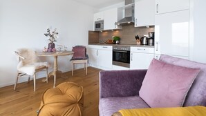 A modern kitchen with white cabinets, a built-in oven, and a microwave. There is a dining area with a round table and two chairs, one with a white fur throw. A purple sofa with a yellow throw blanket is in the foreground.
