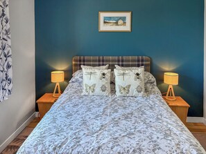 A bedroom with a bed, two bedside tables, a nightstand, a lamp, a painting on the wall, and a floral curtain.
